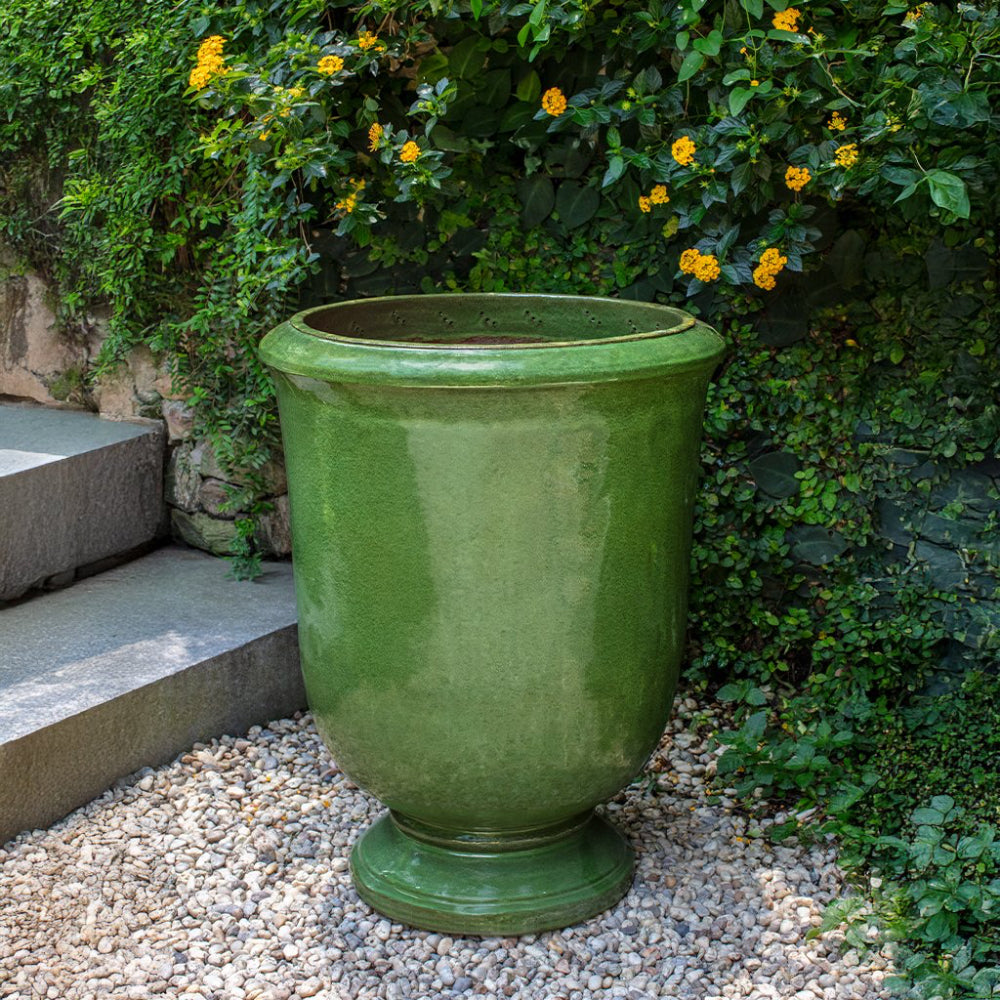 Green ceramic urn in a garden setting with greenery and yellow flowers.
