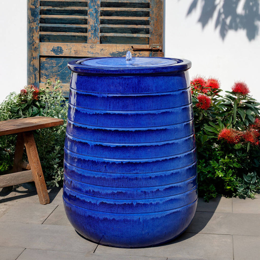 A large ceramic rain barrel fountain with a glossy cobalt blue glaze, featuring horizontal ridges and a small water fountain spout on top. It is set outdoors on a stone patio next to a wooden bench and green plants with red flowers, in front of a weathered blue wooden shutter.