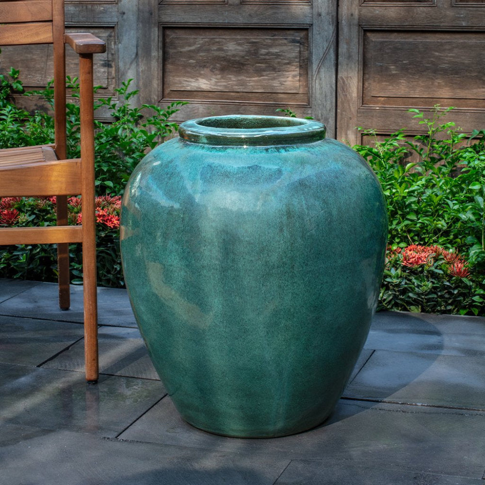 Large teal ceramic vase on a patio with a wooden chair and plants in the background