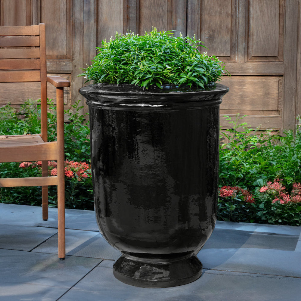 Black urn planter with green plants on a patio with wooden chairs and garden background