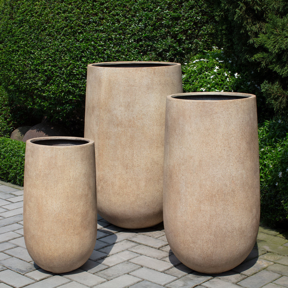 Three beige outdoor fiber lite planters on a stone patio with greenery in the background