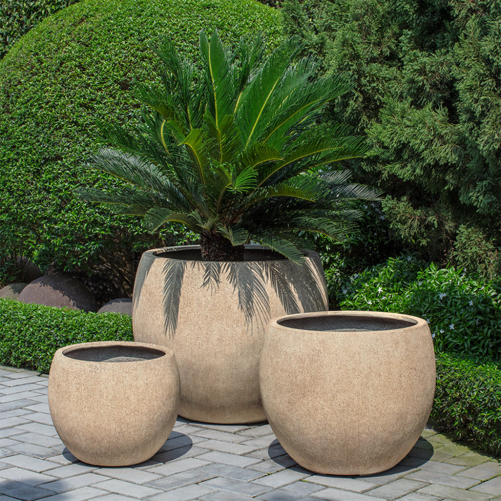Set of three beige planters with a potted plant on a paved area with greenery in the background
