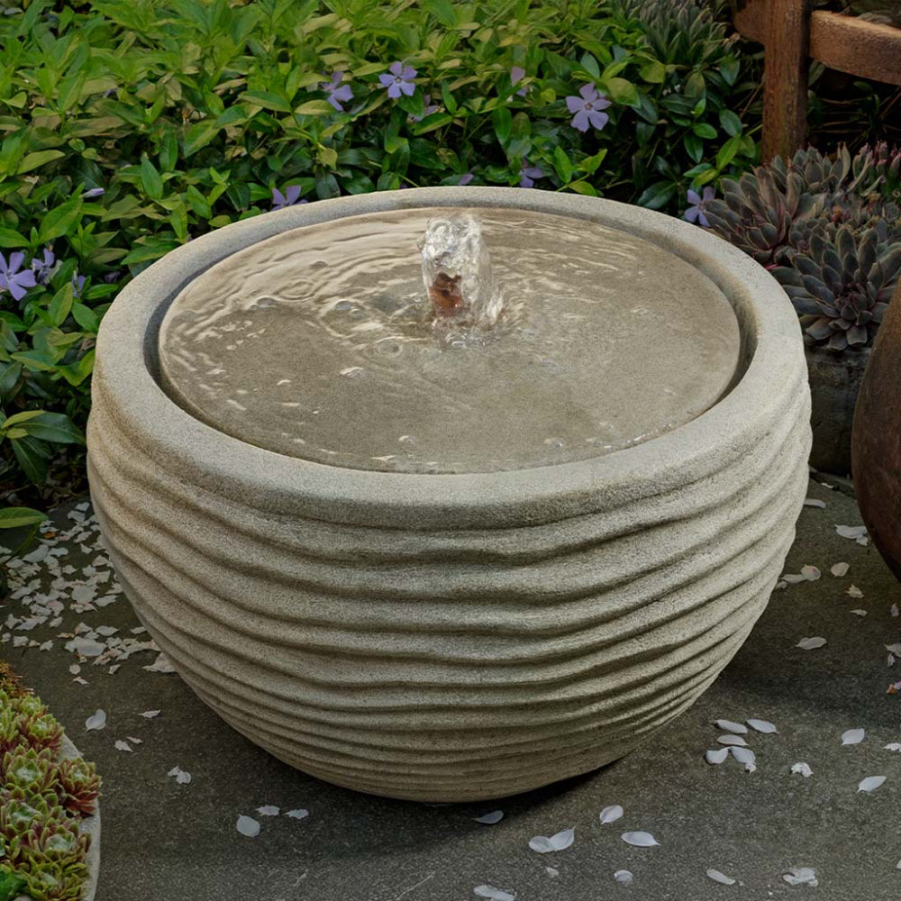 Decorative cast stone water fountain in a garden setting with plants and flowers.