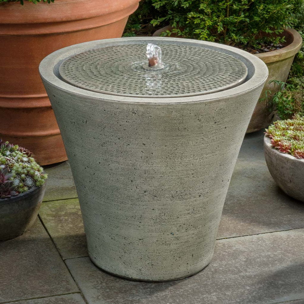 Cast stone outdoor fountain with water feature surrounded by potted plants on a patio.