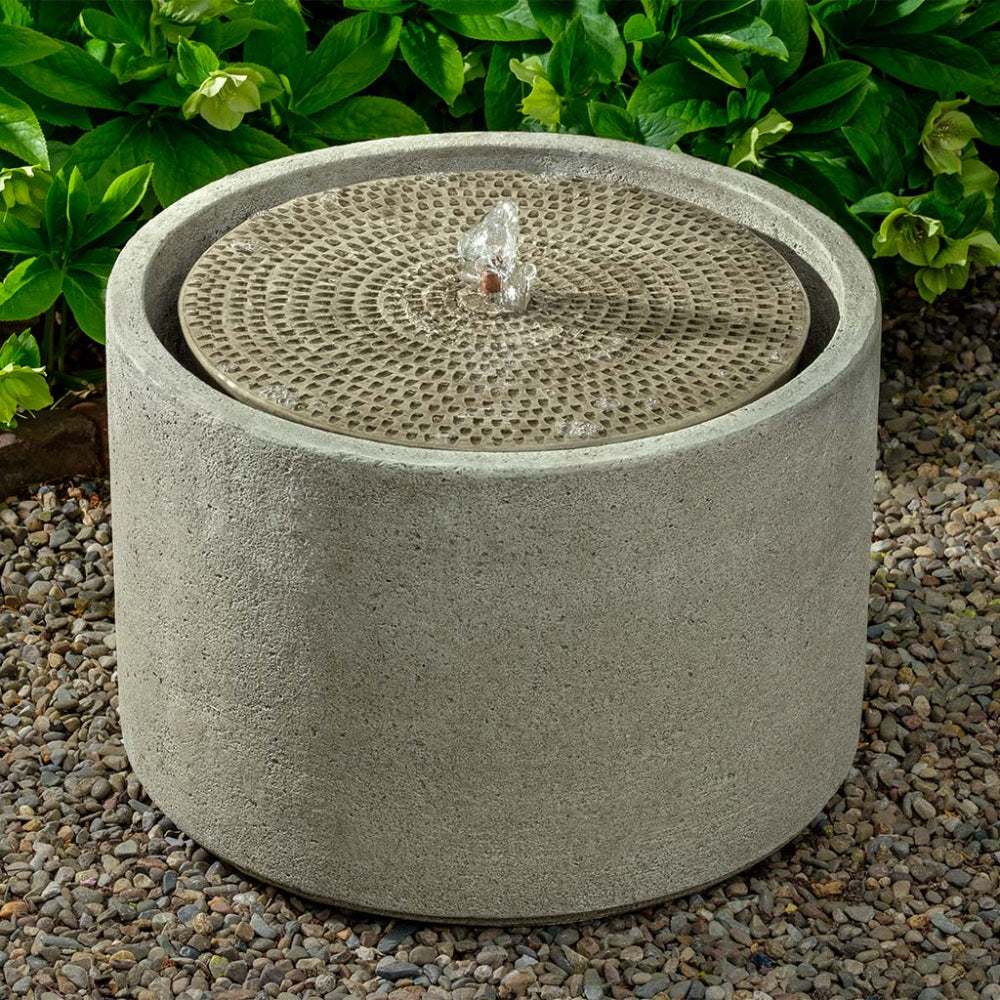 Cast stone water fountain with decorative top on a gravel surface with greenery in the background