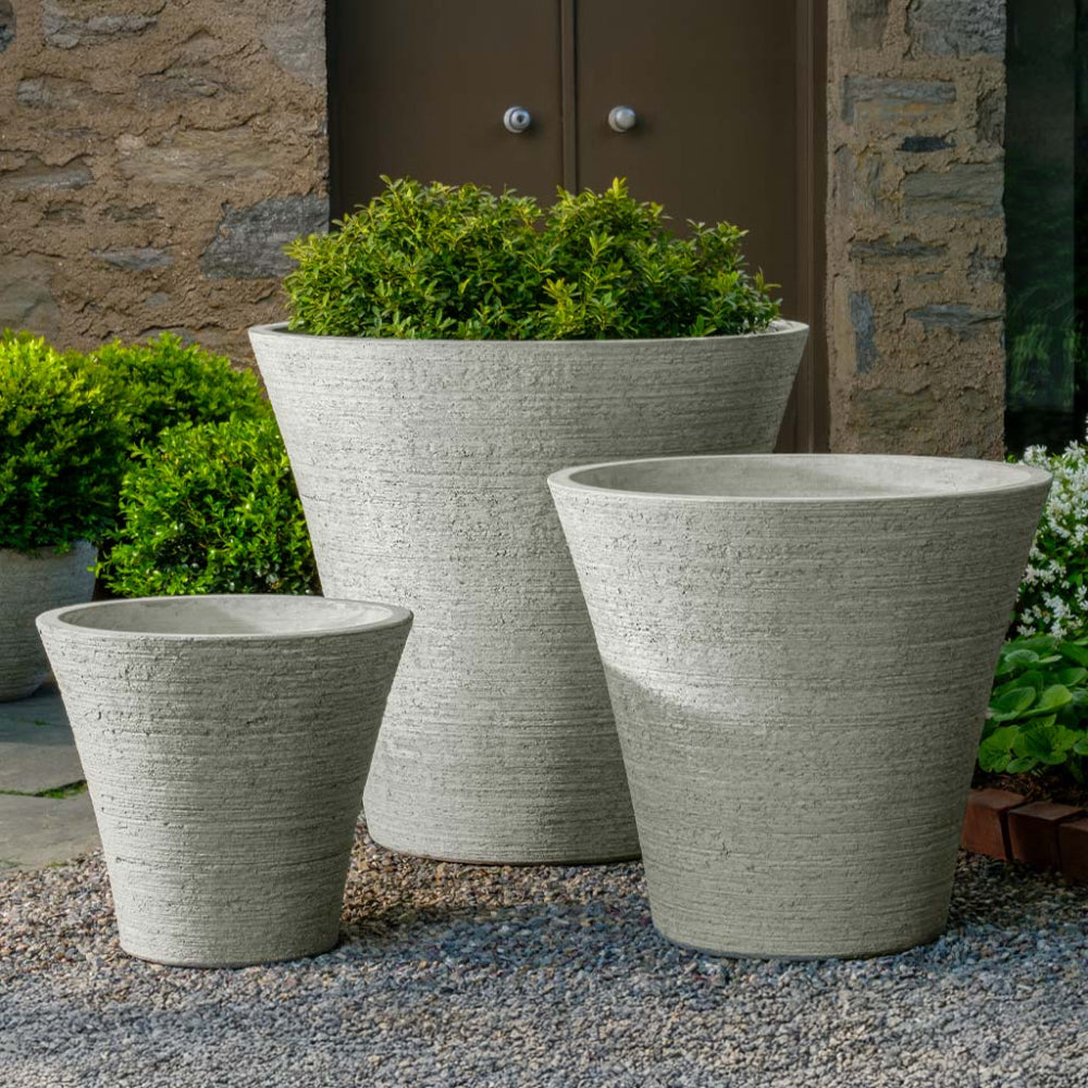 Three stone-textured planters with green plants against a stone wall.