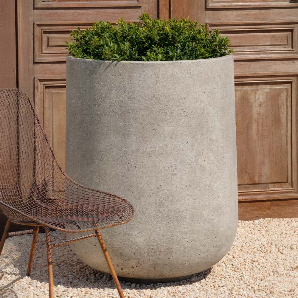 Large cast stone planter with green plants next to a wicker chair against a wooden wall.