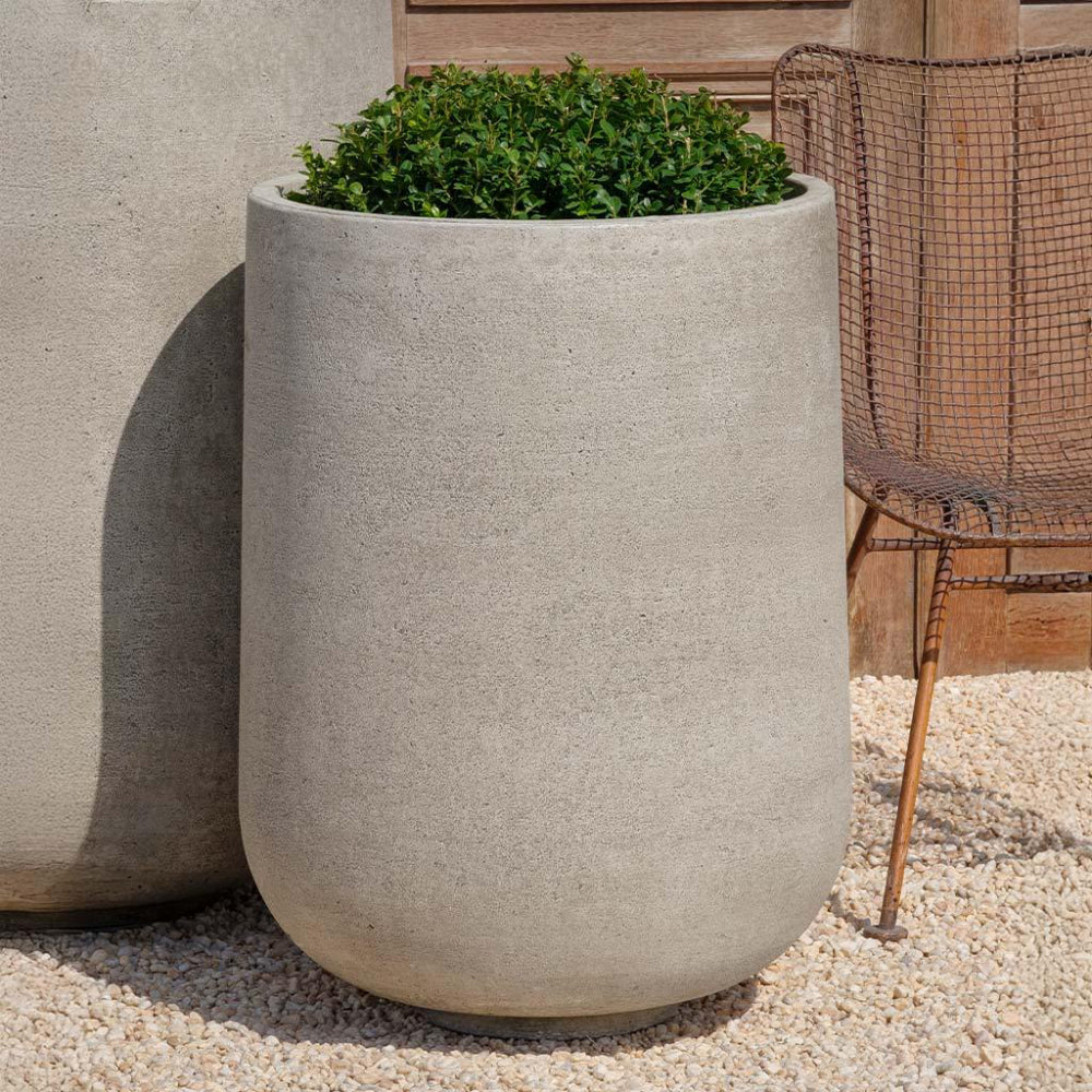 Cast stone planter with greenery on a gravel surface next to a wooden bench.