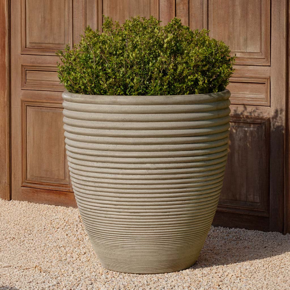 Large ribbed cast stone planter with green plants against a wooden background