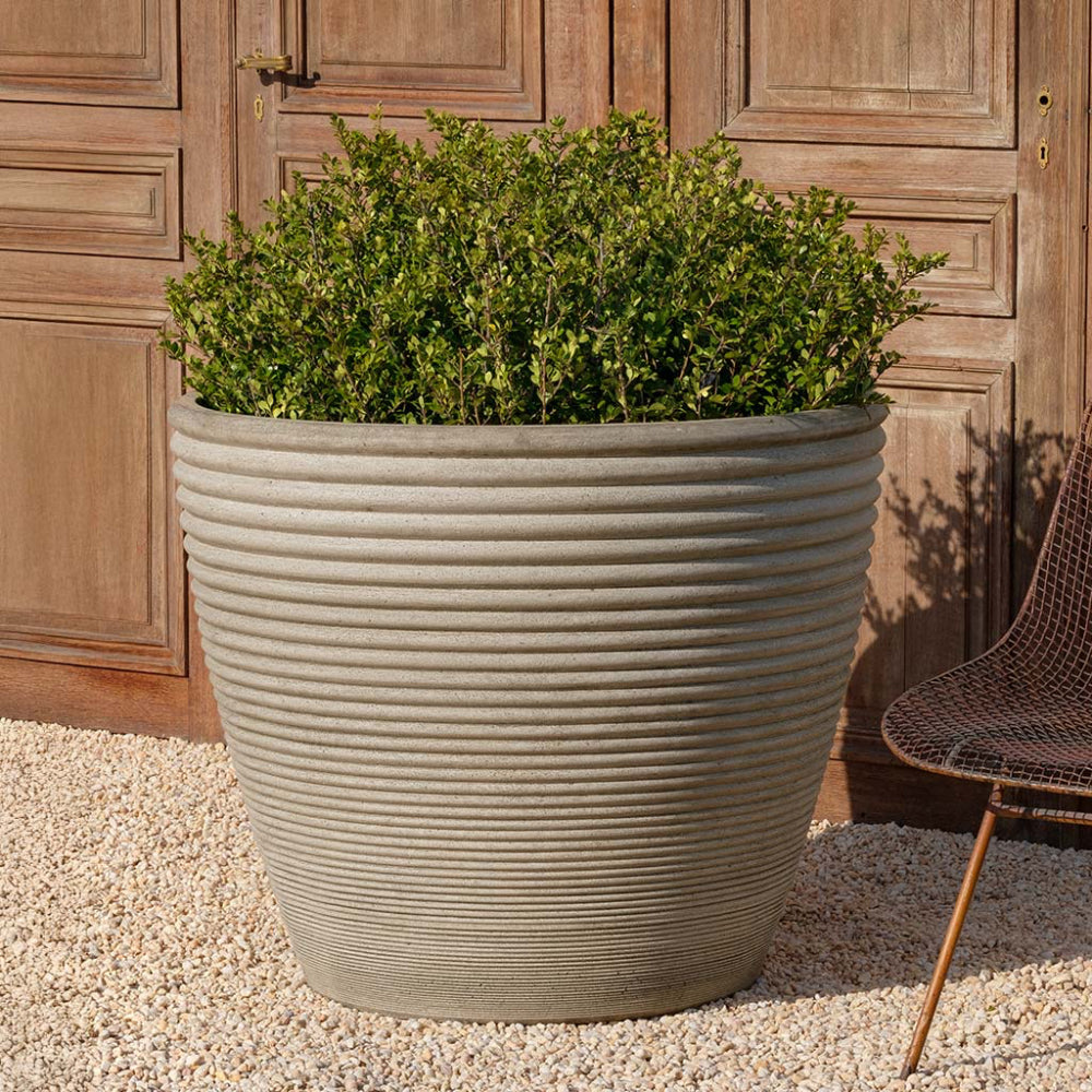 Large ribbed cast stone planter with green plants against a wooden door.