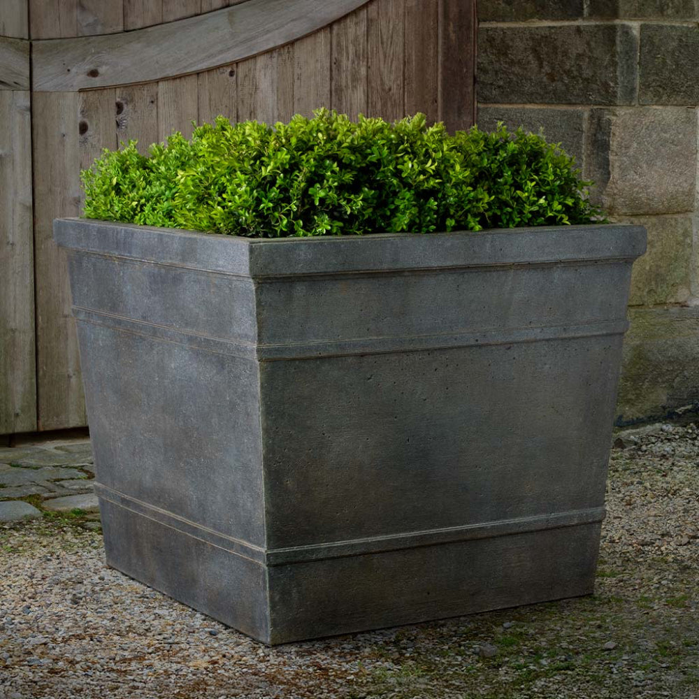 Gray rectangular cast stone planter with green plants against a stone wall.
