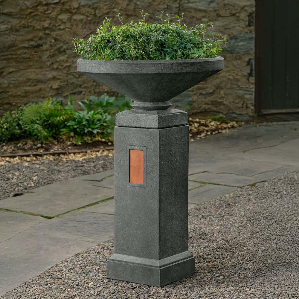 Decorative cast stone urn planter on a pedestal with a copper accent with greenery on a stone patio